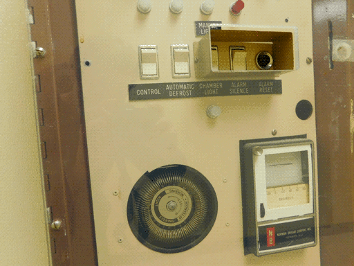 Close up of bottom half of the control panel with the "MANUAL LIGHTS," two push switches labeled "CONTROL" and "AUTOMATIC DEFROST," two more switches and a button (accessible through a slot in the plastic) labeled "CHAMBER LIGHT," "ALARM SILENCE," and "ALARM RESET," a white light underneath the "CHAMBER LIGHT" label, some kind of coil with a labeled central plate, and a thermostat.