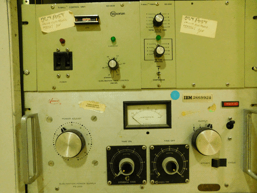 Two modules, the top painted a pale maybe-green-maybe-badly-lit-grey and the bottom a white or pale grey. The top is a Varian Ti-BALL CONTROL UNIT with a Ti-BALL TIMER. The panel has a breaker-style on switch, green "CURRENT" light, silver "SIMULATION RATE CONTROL" knob, and linear "LIFE" meter. The TIMER sub-panel has two silver knobs, "TOTAL CYCLE TIME (HOURS)" and "ON TIME (MINUTES)," a green light, and a toggle switch between "TIME" and "CONTINUOUS". There are two notes pasted on the module, both reading "OUTPOST / VACUUM ELECTRONICS / FRAGILE! / 10F. The bottom module is lablled Veeco and "IBM 2669924", and is a PS-202 SUBLIMATION POWER SUPPLY. It has two larger silver knobs ("POWER ADJUST" and "OUTPUT", two smaller knobs on base plates, a red "POWER ON" button, a push switch between "STEADY" and "CYCLE," handles, and an AC ammeter.