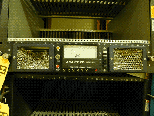 Mostly empty chassis with what looks like a power supply, black, mounted across the middle. There's a rectangular meter in the center measuring no visible units, but labeled "J. WHITE CO. SERIES 800 POWERED CAMAC CRATE." Underneath are 6, 12, and 24 volt sockets (2 of each, positive and negative). To the left are two lights ("ON" and "HI TEMP") and a power switch; to the right are five rectangular buttons. To either side are large vents, with slightly smashed-in multilayer metal filters. The top edge of the module is labeled with numbers 1-25, as is the top edge of a hollow unit directly underneath this module.