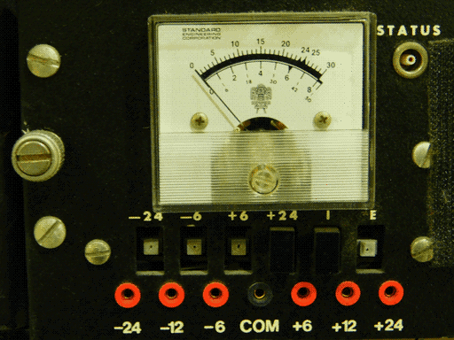 Close-up of a square STANDARD ENGINEERING CORPORATION meter on black plate- presumably a voltmeter. Above the marks, the meter has a 0-30 scale on fives, with 24 specifically present as well, and below the marks, a 0 to tad-over-8 and a smaller 0-50 scale. The meter also has a Mesoamerican looking logo. To the right of the meter is a STATUS port, and below are rectangular buttons to set the voltage to -24, -6, +6, and +24, and two more labeled I and E. All but the +24 and I buttons are missing their caps. Below the buttons are ports labeled -24, -12, -6, COM, +6, +12, and +24, the COM port black and the rest red.