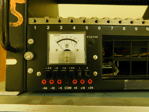 Zooming out from the another of the same meter a little, we see it is on the left side of a rectangular unit similar to the last described. This one has no missing button caps and a blocked off STATUS port. The top edge is numbered, with only 1-20 visible. To the right of the meter is part of a rectangular gap, with a wide grid and some wire bundles inside. Below the gap is a velcro strip. The whole unit appears to be in a chassis below an empty section. To its left on the black chassis frame is a handle.