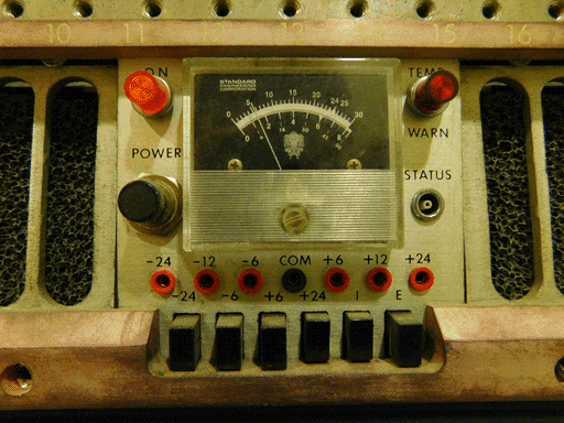 A close-up view of another assembly, with an unusually elegant appearance. Its panel surface is brushed grey metal, with a thinner top bar and thicker bottom bar of a pale copper color. The visible part of the top bar is numbered 10-16. At the center of the assembly is the actual control panel, with another STANDARD ENGINEERING CORPORATION meter with same logo and scales, but printed white on black. To its left are a red ON light and a black POWER button, and to its left are a red TEMP / WARN night and a STATUS socket. Below are seven sockets and 6 buttons, also same as on the last panel (no button caps are missing). Half the width of the bottom bar is cut out around the buttons. To either side of the central control panel are plates with holes cut out (rectangles with semicircle ends), beneath which is a foam filter material. Above the module is a metal bar with regularly spaced screw holes.