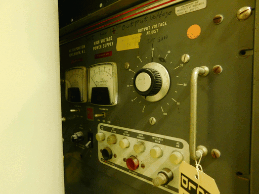 A dark grey control panel under yellow light, its left side receding into darkness behind a corner. It is the face of a HIGH VOLTAGE CONTROL PANEL manufactured by NJE CORPORATION of KENILWORTH, N.J. Its controls are all black, grey, white, or silver, and consist of buttons, a large knob, and toggle switches. It has two NJE meters, rounded squares, measuring D-C kilovolts and D-C milliamps. Along the top of its face is written "Set for (+) Output Voltage" in marker. On its right side it has a silver handle with a white HOLD tag tied to it.