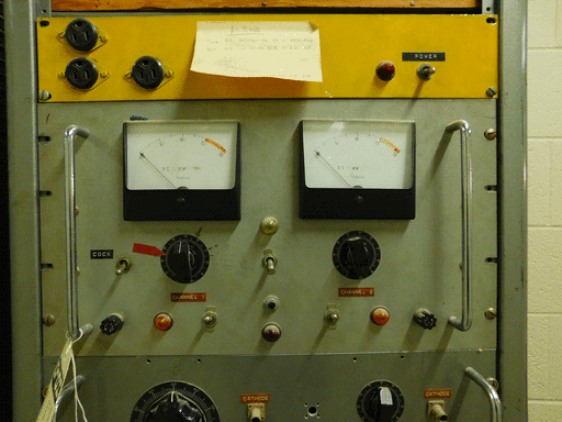 Image centered on a grey control panel with two metal handles. Below it is the top of another panel, slightly darker grey, and above it is a yellow panel. The yellow panel has three 3-prong power sockets, a power toggle and light, and a wide sticky note titled X-BAND with instructions for using the controls below. The central control panel has two Simpson meters measuring DC kilovolts on a 0-20 scale. Below each meter is a black knob, respectively labeled CHANNEL 1 and CHANNEL 2, a fuse, an orange light, and a toggle. Between the two meters' associated controls is a column consisting of: a white light, a toggle, a button, and a red light. There's also a toggle switch under the left meter labeled COCK, and what appears to be a HOLD tag on the left handle. There's visible dust on the lights. This thing hasn't been touched in a while.