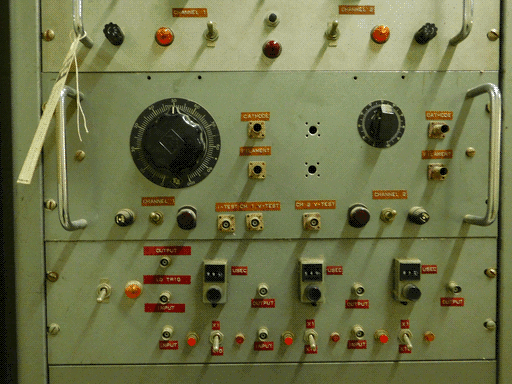 Image centered on the aforementioned lower, slighly darker grey panel, and another panel (slightly shorter) below it. The upper panel of these two has seven sockets, two toggle switches, two red lights, two fuses, and two knobs. One knob is larger and nine-pointed, and one is smaller and linear rather than radial in shape. The controls have label strips with a wood-grain texture and white text, and there are metal handles on either side of the panel. The lower panel of the two has eight sockets, four toggle switches, two small red lights, one large orange light, three bright red buttons, and three rectangular assemblies each labeled USEC. These assemblies' upper halves have 3-digit analog displays like a combination lock, and their lower halves have black knobs. They are set to 997, 549, and... either 876.5 or 877.5. These controls have red label strips with white text.