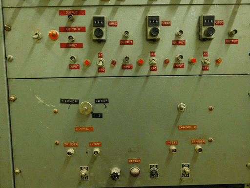 This picture shows the lower panel from the previous one, and (surprise) another taller panel below that. It's got two toggles, four sockets, a very dusty large red light, and a green on/off button. Actually two of those toggles are also on plates labeled "ON OFF". I'm a little confused. There's also a screw on a plate with three white-on-black labels: E.3 below, HIGHER to the left and LOWER to the right, the latter two above sharpied CCW and CW arrows. I guess you needed a screwdriver to use this control. (There have been other such screws on previous control panels I neglected to mention out of befuddlement.) Anyway the rest of the control labels are white on woodgrain again.