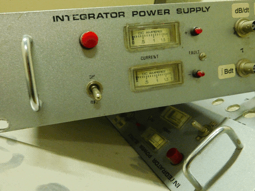 Two removed modules, only front plates visible, one leaning atop the other. Each is a long rectangular "INTEGRATOR POWER SUPPLY" with speckled metal finish and a metal handle on each side. They each have a large red (power?) light and an on/off toggle switch. They also have two current meters reading "DC AMPERES", one also labeled with a plus and one with a minus sign. These have linear rather than radial displays, but they are either printed on the edges of rotating discs recessed in the panel or are printed on spools of tape. Each has a smaller red light next to it. There are then two sockets labeled "dB/dt" and ∫Bdt.