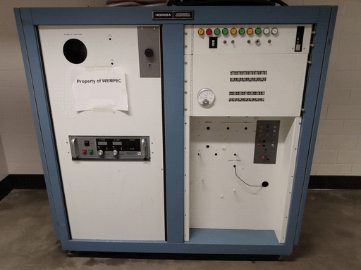 A large assembly of control panels, appearing largely bare for its size, white with a dull blue frame diving it into halves vertically. At the top of the frame is a nameplate reading "HORIBA INSTRUMENTS INCORPORATED." The left control panel has a large round hole labeled "SAMPLE VENTURI", two small ones labeled "HIGH ALARM" and "LOW ALARM," a metal plate with an octagonal knob and a toggle switch, a piece of paper taped on reading "PROPERTY OF WEMPEC," and a power supply. The right side has a control panel in its top half and a recessed portion in its bottom.