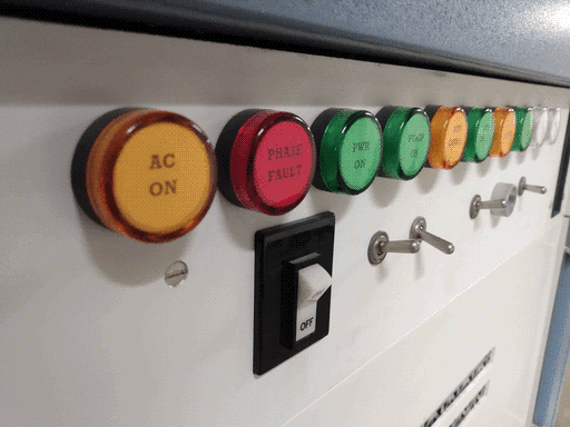 Close-up of 10 indicator lights on white backing, left side closer to the camera: orange, red, green, green, orange, green, orange, green, white, white. They are labeled "AC ON," "PHASE FAULT," "PWR ON," "PUMP ON," and "BUS CONN." and then they get too blurry to read. Below them there's an off switch and some toggle switches